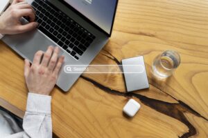 Persona utilizando un ordenador portátil en un escritorio de madera con una barra de búsqueda digital, representando la optimización SEO y el diseño web estratégico.